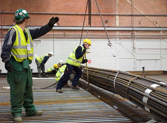 Workers in safety vests and hard hats are installing a large steel pipe at a construction site for carpet reinforcement